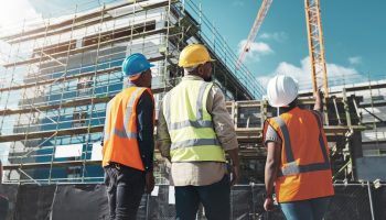 Three People In Hi-vis Vests And Hardhats Stand Just Outside A Black Construction Fence Around The Base Of A Large Commercial Building Being Built.