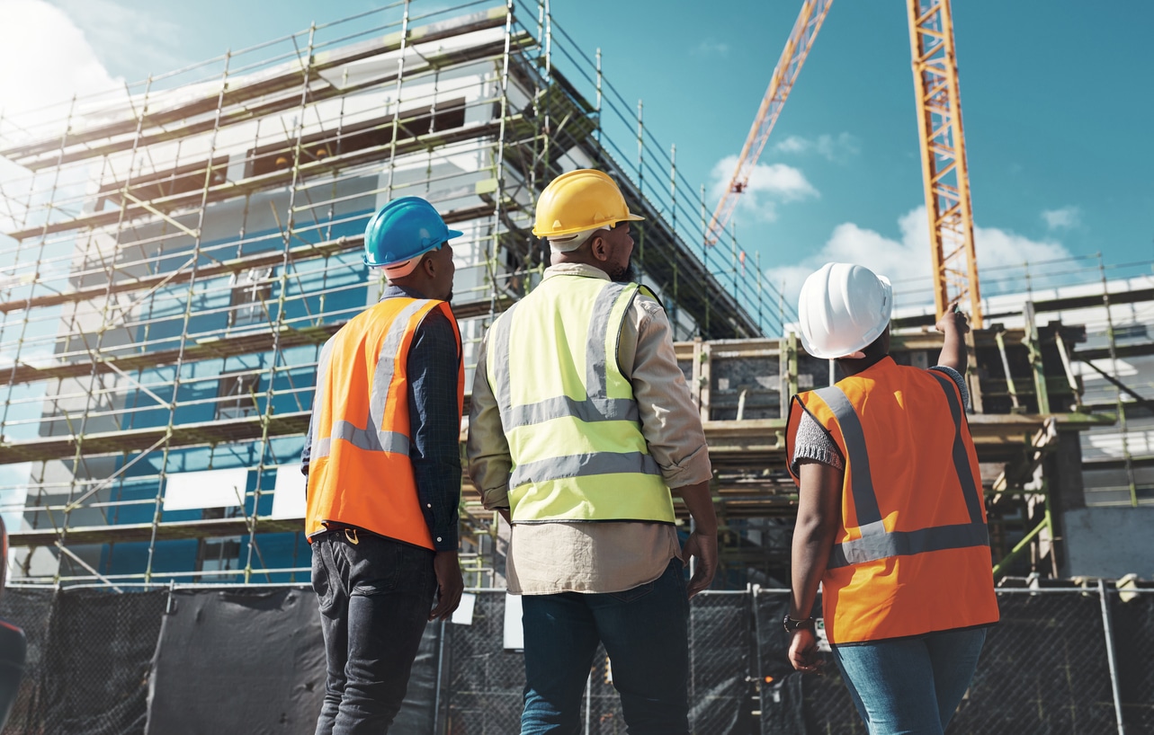 Three people in hi-vis vests and hardhats stand just outside a black construction fence around the base of a large commercial building being built.