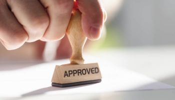 An Image Of Someone Stamping A Paper With A Stamp That Reads “Approved”.