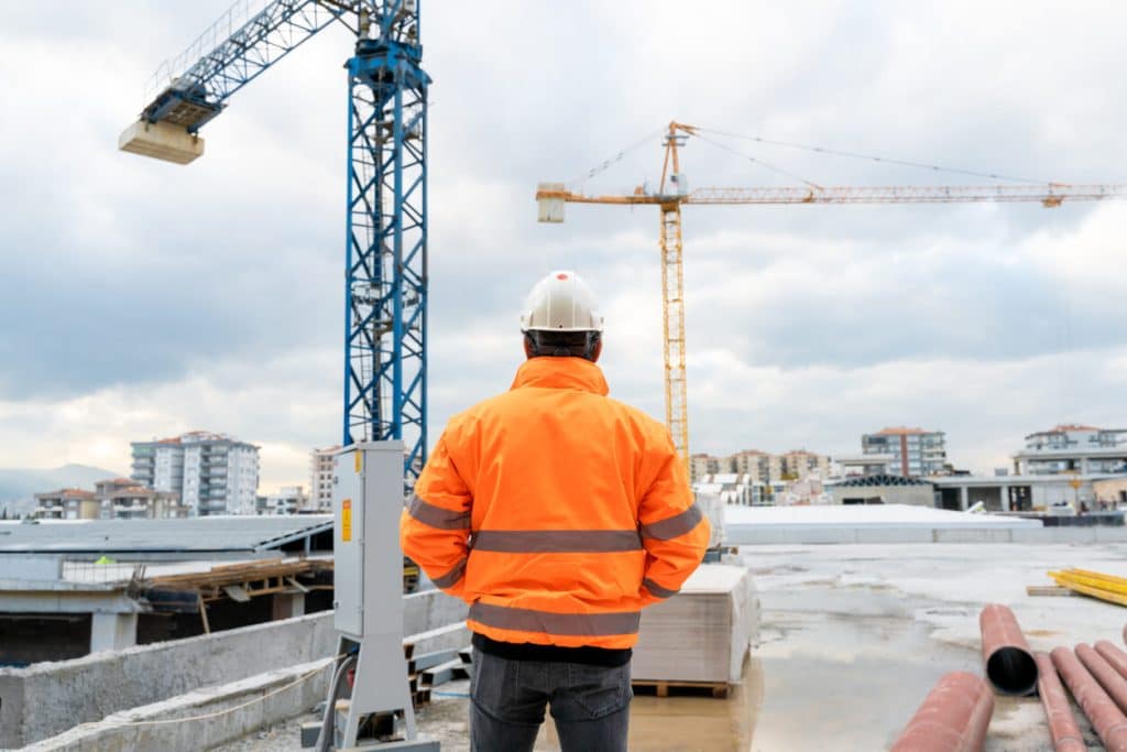 A man in a hard hat and orange vest examines work at commercial building under construction.