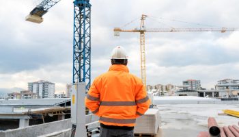 A Man In A Hard Hat And Orange Vest Examines Work At Commercial Building Under Construction.