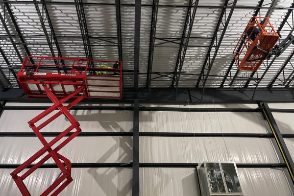 Construction workers installing steel framing and roof systems inside an industrial building using aerial lifts.