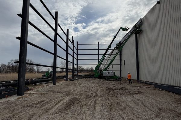 Steel frame addition under construction beside metal warehouse with boom lifts and worker on gravel site